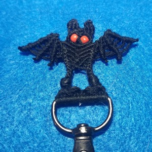A keyring with dark metal with a black bat with red eyes hanging on it by its feet, photographed on a blue background