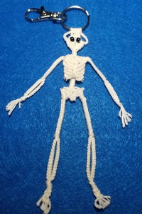 A keyring with silver metal with a white skeleton with black eyes hanging on it by its head, photographed on a blue background