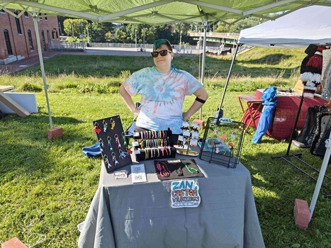 A person standing behind a table at an arts festival. The table has dozens of colorful weavings on display.